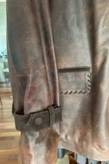 Tapado de cuero marrón con diseño artesanal, cuello camisero con detalles de pasamanería en gamuza, cierre frontal con botones de guampa y bolsillos delanteros con bordes decorativos. Presenta un corte cómodo y versátil.