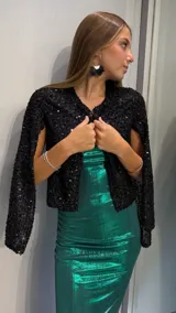 Chaqueta corta de color negro, confeccionada en tejido cubierto de lentejuelas brillantes. Presenta un diseño abierto sin cierre frontal, con mangas tipo capa que aportan un estilo elegante y sofisticado.