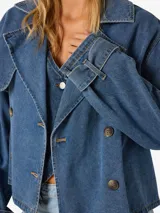 Trench corta de denim azul claro, con cuello solapa estilo biker, doble botonadura frontal y diseño holgado.