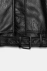 Cazadora de cuero sintético negro con textura granulada, cuello alto, manga larga y cierre frontal con cremallera oculta. Cuenta con bolsillos delanteros y un cinturón ajustable con hebilla metálica en el bajo.