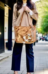 Cartera tipo shopper de cuero suave color marrón terra, con diseño pintado a mano que incluye salpicaduras y la inscripción de la marca. Cuenta con dos asas largas para llevar al hombro, cierre principal, forro interior con bolsillo y un dije metálico colgante.