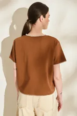 Remera marrón de manga corta y cuello redondo, con detalles de costuras decorativas en contraste color crudo en el cuello, los puños y el dobladillo inferior. Cuenta con el nombre de la marca impreso en el pecho.