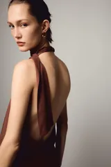 Vestido midi de color marrón con cuello halter y lazada en la nuca. Presenta un diseño con espalda descubierta, frunces en la cintura y una abertura lateral en la falda.