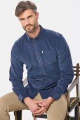 Camisa de pana fina en color verde oscuro, de manga larga con puños arremangados, cuello camisero con botones y un bolsillo aplicado en el pecho con pequeña etiqueta decorativa.