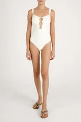 Malla enteriza de color crema con diseño strapless, fruncidos en el frente y tres aberturas circulares verticales separadas por herrajes metálicos brillantes. Presenta tirantes finos removibles.