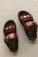 Sandalias bajas de cuero marrón con doble tira, hebilla ajustable y apliques de tachas metálicas.
