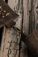 Bandolera confeccionada en cuero de gamuza marrón, con cierre superior. Presenta un llavero colgante con forma de estrella revestido en tachas metálicas y tiras flecadas en tono dorado.