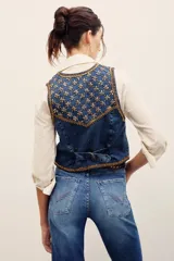 Chaleco de denim azul con bordados metalizados y pedrería en la parte superior delantera, escote en V y cierre frontal con botones metálicos.