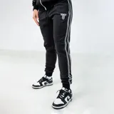 Pantalón de jogging negro con diseño deportivo, presenta tres líneas blancas verticales a lo largo de los laterales y un logo bordado en color blanco en la parte superior del muslo. Cuenta con cintura elástica con cordón ajustable y puños elásticos en los tobillos.