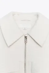 Cazadora de textura suave con cuello solapa, manga larga y cierre frontal con cremallera metálica. Presenta dos bolsillos de plastrón con solapa en la parte delantera.