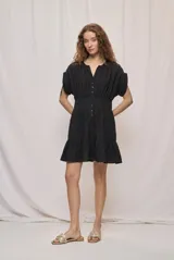 Vestido corto negro confeccionado en algodón, con silueta marcada en la cintura y falda con volumen. Presenta escote redondo con abertura, botones frontales, mangas cortas amplias y terminación con volado en el ruedo.