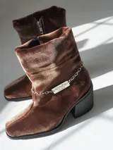 Bota corta de velvet azul marino con efecto abullonado en el tobillo y cierre lateral con cremallera. Tiene un tacón cuadrado y una cadena desmontable con el logo de la marca.