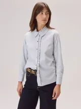 Camisa de algodón color gris claro, con cuello clásico y detalle de volados en la cartera frontal. Presenta mangas largas con puños abotonados y un corte recto.