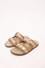 Sandalias bajas color beige con doble tira, una de cuero color tostado y otra de cuero color beige claro con hebilla dorada ajustable. Las tiras están adornadas con tachas doradas.