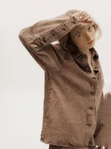 Campera de denim gastado color marrón, de corte holgado y ligero, con cuello camisero, cierre frontal de botones y detalles de botones en los puños.