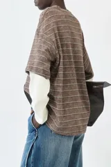 Remera oversize de manga corta superpuesta a una manga larga, ambas de algodón. La capa superior es marrón con rayas horizontales finas en blanco y presenta un efecto lavado. La capa inferior es de color crema o blanco roto.
