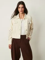 Campera de denim color off white con cuello camisero, bolsillos con solapa en el pecho y cierre frontal con botones metálicos. Presenta detalles de bordado con perforaciones en las mangas.