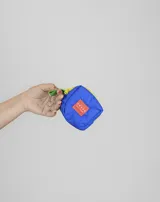 Monedero pequeño de tela color azul, con cierre amarillo y correa de mano verde con gancho metálico. Presenta una etiqueta rectangular naranja con el logo de la marca en el frente.