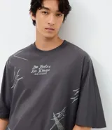 Remera de corte oversized color gris oscuro, con cuello redondo y manga corta. Presenta una estampa gráfica en la espalda que incluye un ángel, estrellas y el lettering 'No Mercy' junto a la frase 'Las calles no perdonan'.