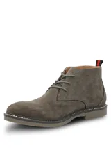 Botines tipo chukka Hush Puppies modelo New Safari, confeccionados en gamuza color gris. Presentan cierre de cordones, suela de goma con costura decorativa y detalle de tirador en el talón con franjas rojas y negras.