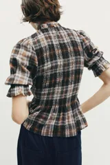 Blusa de cuello subido con diseño de cuadros en tonos marrones, negros y blancos. Presenta mangas cortas con terminación en volantes y un corte ajustado con detalle de volado en la cintura.