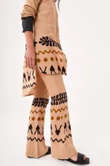 Pantalón de lino color beige, de corte flared y tiro medio alto. Presenta cierre y trabilla metálica, bordado en la pretina con hilo plata y estampado por serigrafía con figuras en marrón y negro.