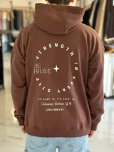 Canguro oversize marrón con capucha, bolsillo tipo canguro y diseño estampado en la espalda con el texto "Be Brave" y detalles tipográficos en color blanco.