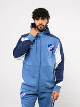 Campera deportiva Umbro del Club Nacional de Football, con cierre frontal, capucha ajustable con cordones y diseño en bloques de color azul, celeste y blanco. Presenta el logo de la marca y el escudo del club bordados en el pecho.