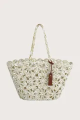 Bolso tipo cesta tejido a crochet con diseño de flores caladas en color blanco y detalles dorados. Cuenta con asas trenzadas y una etiqueta decorativa de cuero sintético marrón.