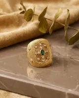 Anillo ancho de bronce con baño de oro, diseño texturizado con detalles calados tipo filigrana, incrustaciones de circonitas y una piedra verde central ovalada.