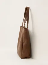 Bolso tipo tote de cuero color marrón, con diseño minimalista, doble asa de mano y cierre superior. Presenta el logo de la marca grabado en un parche central y detalles de remaches metálicos.