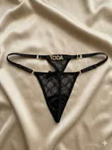 Tanga de tul translúcido negro con diseño de encaje tipo monograma, tiras elásticas regulables con argollas doradas y detalle de letras en strass formando la palabra "TODA".