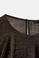 Top de cuello redondo y manga sisa, confeccionado en tejido con acabado satinado. Presenta volantes en cascada y un diseño de bajo asimétrico, con cierre en la espalda mediante pequeña abertura y botón.