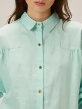 Camisa de lino color verde agua, con cuello clásico, cierre frontal de botones y mangas largas con puños. Presenta detalles de costuras en zigzag en la zona de los hombros y canesú.