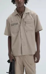 Camisa de manga corta color beige, estilo técnico, confeccionada en microfibra liviana. Cuenta con doble bolsillo frontal, uno con detalle de malla y otro con cierre, y broches en lugar de botones.