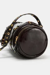 Bolso estilo bowling de cuero sintético acharolado color marrón, con diseño cilíndrico, bolsillos laterales con cierre y doble asa decorada con tachas metálicas doradas.