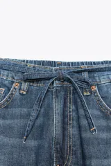 Pantalón de denim azul de corte amplio, con cintura ajustable con cordones, bolsillos laterales y traseros, y cierre frontal con cremallera y botón metálico.