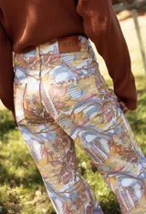 Pantalón tipo jean de corte recto con estampa exclusiva de diseño artístico en tonos tierra, ocres y azules. Presenta bolsillos traseros y cierre con botón metálico.