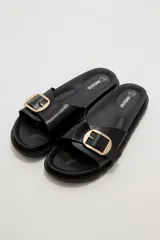 Sandalia plana de eco cuero negro, con tira ancha en el empeine decorada con una hebilla metálica dorada cuadrada y suela dentada.