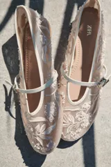 Bailarina de mesh color beige con bordados florales y detalles de perlas aplicadas. Presenta una tira delantera con hebilla metálica y acabado en punta redonda.