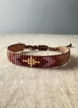 Pulsera tejida con cuentas Miyuki en tonos tierra, granate y rosa, con un diseño central geométrico en color dorado. Cuenta con un sistema de cierre ajustable mediante cordón marrón.
