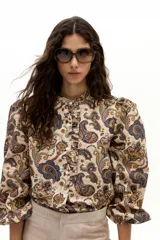 Camisa de algodón con estampado paisley en tonos beige, azul y naranja. Presenta cuello mao, hombros abullonados, mangas largas con puño elástico y botones de nácar en el frente con un detalle de vuelo.