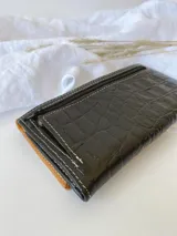 Billetera de cuero con diseño tricolor en tonos verde, marrón y negro, con textura tipo reptil. Presenta un parche central con la marca grabada y cierre con broche.