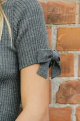 Jersey de punto acanalado en color gris, con cuello redondo y mangas cortas que presentan detalles de lazos ajustables en los extremos.