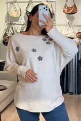Buzo tejido de punto en color beige, con diseño de flores aplicadas en tono gris oscuro y detalles de brillos plateados que irradian desde las flores. Presenta cuello redondo amplio, mangas largas con caída holgada y corte recto.