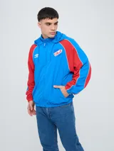 Campera rompevientos Umbro del Club Nacional de Football, modelo Venti para hombre. Diseño en color azul con mangas rojas y detalles blancos en los recortes, capucha con cordón ajustable y bolsillos laterales. Cuenta con el logo de Umbro y el escudo del club bordados en el pecho.