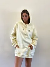 Hoodie oversize color beige con capucha y cordones ajustables. Presenta un pequeño logo bordado de un león con corona en el pecho y un bolsillo frontal tipo canguro.