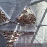 Conjunto de lencería de dos piezas confeccionado en tela acanalada blanca con estampado de pequeñas flores rosas. Incluye un soutien tipo triángulo sin aro, con detalles de encaje blanco en los bordes y una argolla metálica en el centro, y una bombacha colaless ajustable con tiras laterales y detalles de encaje.