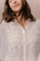 Camisa beige de calce relajado y tejido semitransparente, con delicados bordados florales en relieve, cuello camisero, abotonadura frontal y mangas amplias.