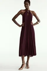 Vestido midi plisado color bordó, con cuello halter, broche de flor en el delantero y anudado en la espalda. Cuenta con forro interior.